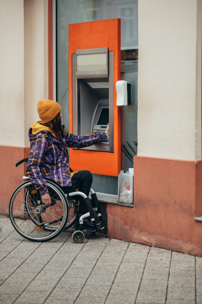 Young beautiful woman with disability who uses a wheelchair using credit card and atm machine while withdrawing money